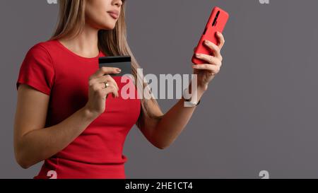 Faceless woman holding credit card and smartphone while making electronic payment. Contactless payment. Faceless Girl in red T-shirt with debit card and mobile. Online shopping on gray background Stock Photo