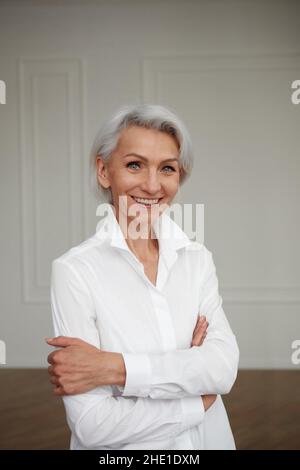 Portrait of an adult elderly self-confident woman with white hair looking at camera in white shirt hands crossed background wall Stock Photo