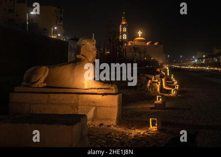 A sphinx on the avenue of the sphinxes in Luxor, Egypt with St. Marys coptic church in the background. Both are illuminated with lights are night. Stock Photo