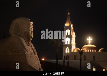 A sphinx on the avenue of the sphinxes in Luxor, Egypt with St. Marys coptic church in the background. Both are illuminated with lights are night. Stock Photo