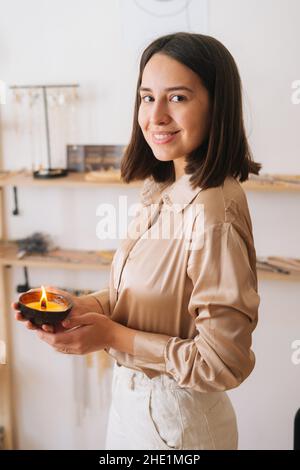 Vertical portrait of smiling young woman holding burning scented handmade candle in palms, looking at camera. Stock Photo