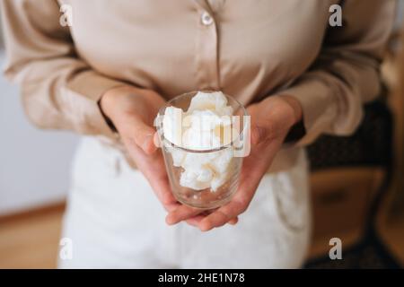 Close-up cropped shot of unrecognizable female artisan holding transparent glass of dry soy wax for creating candle building mixture. Stock Photo