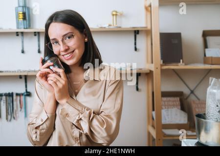 Portrait of cheerful young woman in glasses holding little empty transparent glass jar in hands sitting at table, looking at camera. Stock Photo