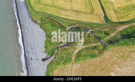 Aerial image of a winding stream running to the sea, you can see fields, beach and sea.  Coastal path goes over the stream via a large slate slab. Stock Photo