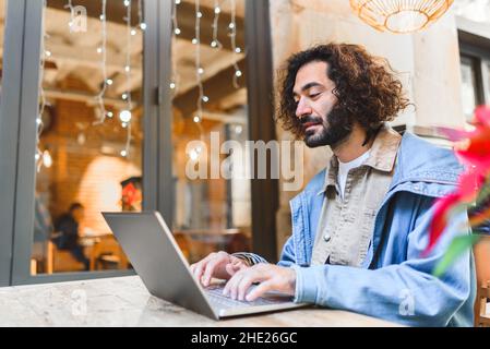 Bearded Hispanic male in casual outfit with dark curly hair. He is typing on laptop keyboard while sitting at table outside cafeteria and working remotely on city street Stock Photo