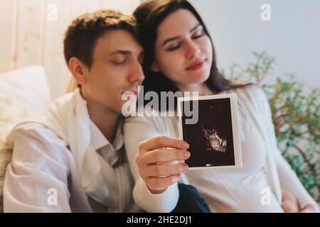 Young happy pregnant woman holding pregnancy ultrasound, showing sonogram picture to pleasantly surprised husband crying from happiness. Family couple Stock Photo