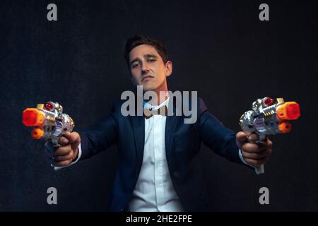 Portrait of serious businessman standing with toy guns against gray background. Stock Photo