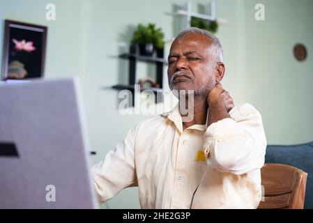 Indian senior old man suffering from neck pain while working on laptop at home - concept of unhealthy sitting posture, arthritis, and joint pain Stock Photo
