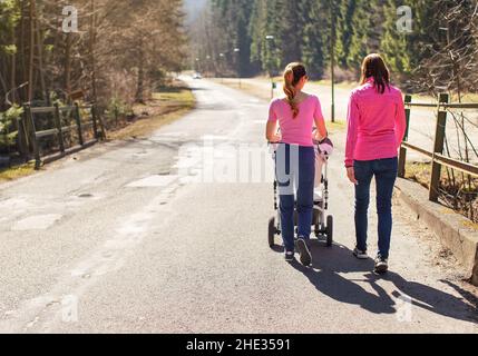 Two young woman in pink tops walking with baby carriage over asphalt road trees both sides, on sunny day, view from behind Stock Photo