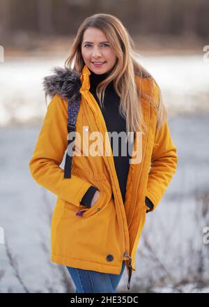 Casual portrait of young woman in warm orange yellow winter jacked, zipper down, smiling, blurred background behind her Stock Photo