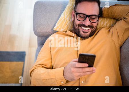 Cheerful young adult mature man happy smiling using smart phone resting lying down on sofa couch at home reading messages on mobile phone Guy with eye Stock Photo
