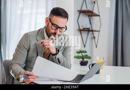 Serious pensive thoughtful focused young adult casual businessman in office working with laptop computer papers and documents looking at paper report Stock Photo