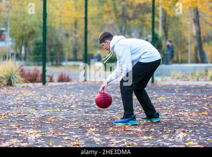Cute teenager in white hoodie playing basketball. Young boy with red ball learning dribble and shooting on the city court. Hobby for kids, active life Stock Photo