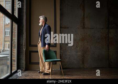 Full length shot of pensive grey haired middle aged man in casual wear looking out a large window while standing in modern loft interior Stock Photo