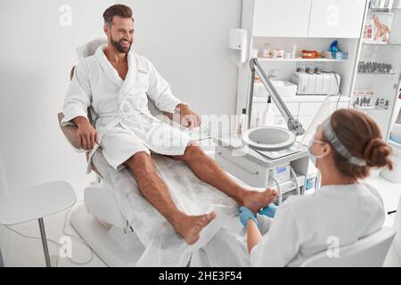 Positive client sits in armchiar while master checks his feet before pedicure in salon Stock Photo