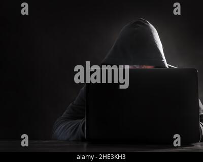 Hooded hacker sitting behind laptop against dark background Stock Photo