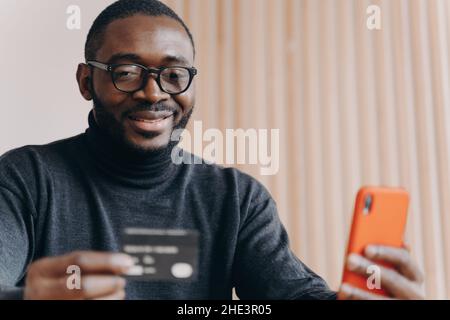 Young positive African ethnicity man entrepreneur in glasses paying with credit card online Stock Photo