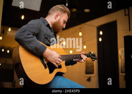 A young guy with a beard plays an acoustic guitar in a room with warm lighting Stock Photo