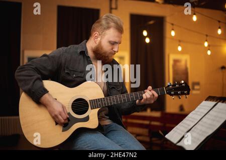 A young guy with a beard plays an acoustic guitar in a room with warm lighting Stock Photo