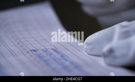 Side view of a hand making notes in a logbook. Close up of hands in white gloves writing data in the notebook with a pen on black background. Stock Photo