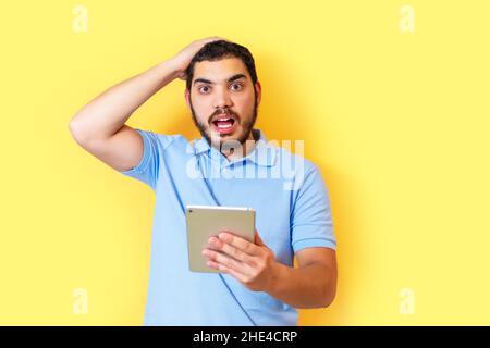 Casual man with beard over yellow wall surprised and holding tablet. Stock Photo