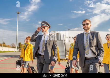 Male pilots and female flight attendants in aviation uniforms walking under blue sky at airport Stock Photo