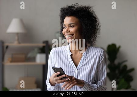 Dreamy cheerful young african woman holding smartphone in hands. Stock Photo
