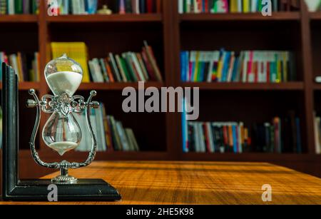 Chiang Rai,Thailand - Sep 06, 2020 : Hourglass on desk with the background of bookshelves in library. Past moments concept with copy space, Selective Stock Photo