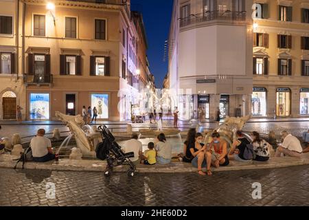 Italy, city of Rome, people relax at the Barcaccia Fountain on Piazza di Spagna by night with view towards Via dei Condotti street. Stock Photo