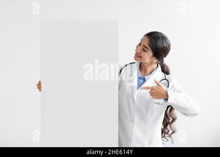 Happy millennial cute hindu lady doctor in uniform shows finger to big banner with empty space Stock Photo
