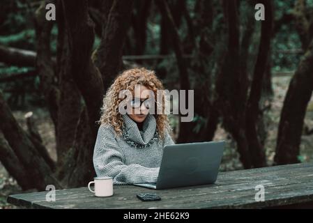Curly long hair blonde adult woman use laptop computer outdoors sitting in the woods - concept of modern free female peoplelifestyle in smart and remo Stock Photo