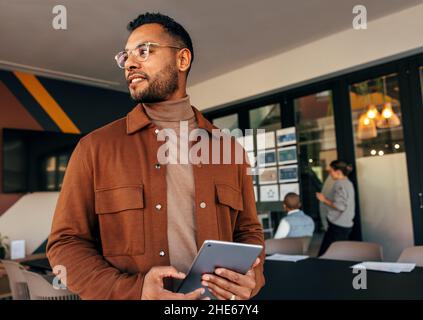 Thoughtful businessman holding a digital tablet and looking away. Contemplative young businessman standing in a modern boardroom with his colleagues i Stock Photo