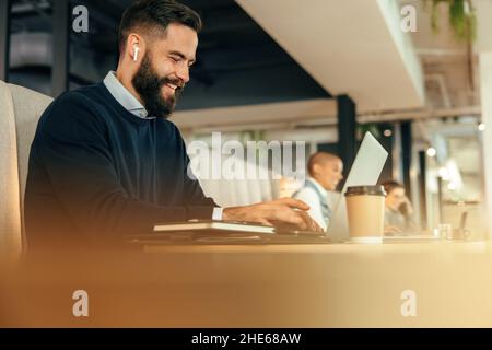 Cheerful young businessman working on his laptop in a co-working space. Modern businessman smiling while typing on his laptop and listening to music. Stock Photo