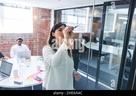 Creative young businesswoman writing on a glass wall with her colleagues in the background. Focused young businesswoman sharing her ideas with her tea Stock Photo