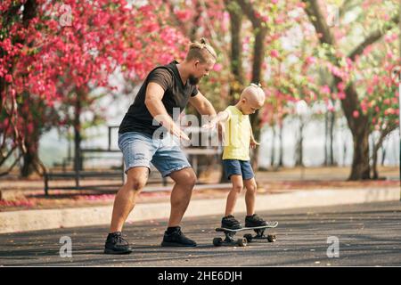 Father holding and training his little son to ride a skateboard in park on spring time. Concept of leisure and outdor family activity. Caucasian man t Stock Photo