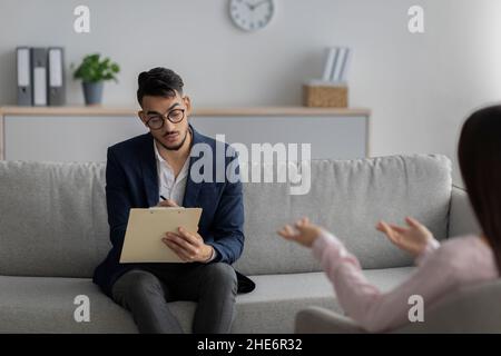 Arab male psychologist talking with female client and taking notes during psychotherapy session, sitting in office Stock Photo