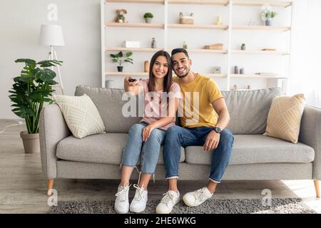 Joyful diverse couple watching TV program, sitting on couch at home, full length portrait Stock Photo