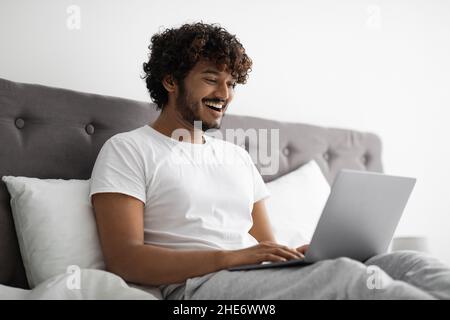 Young indian man typing on laptop, working in bed Stock Photo