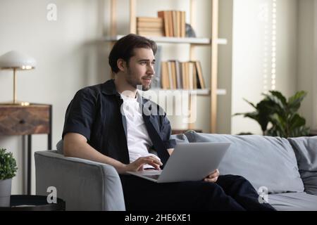 Dreamy smiling young man using computer at home. Stock Photo