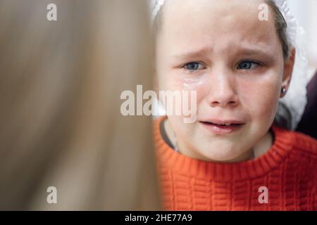 Portrait of little tearful girl in medical disposable cap crying in pain after ear piercing or other medical procedures in hospital office Stock Photo