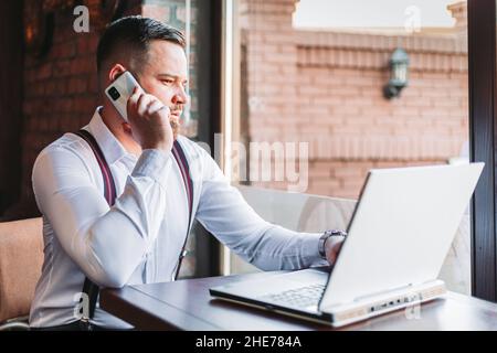Young businessman talking over a phone in a cafe, laptop on the table Stock Photo