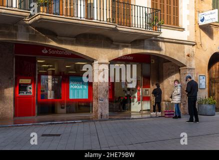 Llucmajor, Spain; january 07 2022: Branch office of Banco Santander with people waiting outside, queuing and wearing masks, in the Mallorcan town of L Stock Photo