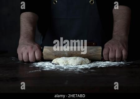 Man Baker or chef is rolling fresh dough with a dough roller on a wooden surface. Stock Photo