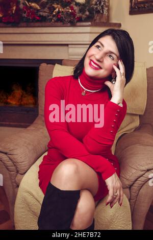 Middle aged brunette woman in the red roll neck dress, sitting in a chair Stock Photo