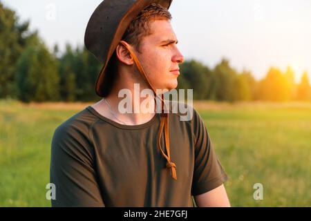 Young man serious farmer in cowboy hat at agricultural field on sunset with sun flare. Profile portrait of man standing on nature background, outdoors Stock Photo