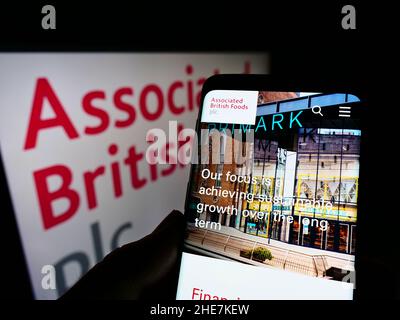 Person holding smartphone with webpage of company Associated British Foods plc (ABF) on screen in front of logo. Focus on center of phone display. Stock Photo