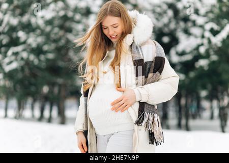 pregnant woman in warm winter clothes standing outside on a snowy winter  day, with hands on her stomach on the background of nature. Concept of  pregna Stock Photo