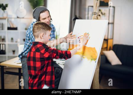 Cute little boy and his caring father using colorful paints for drawing with hands on canvas. Caucasian family of two spending time with creativity at home. Stock Photo