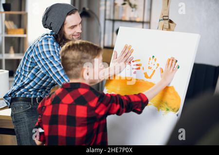 Caring caucasian father teaching his little son drawing on canvas. Handsome man and cute boy in checkered shirts using colorful paints at home. Stock Photo