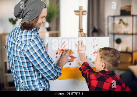 Caring caucasian father teaching his little son drawing on canvas. Handsome man and cute boy in checkered shirts using colorful paints at home. Stock Photo